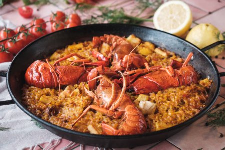 Spanische Meeresfrüchte-Paella mit Hummer und Tintenfisch, traditionelles Gericht mit Reis, Draufsicht auf einen heißen Topf, umgeben von frischen Zutaten auf einem rosa Hintergrundtisch