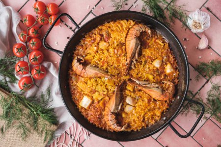 Téléchargez les photos : Paella aux fruits de mer espagnols avec crevettes et calmars, plat traditionnel avec riz dans une casserole chaude, entouré d'ingrédients frais sur une table de fond rose, vue sur le dessus - en image libre de droit
