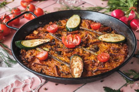Photo for Spanish paella with vegetables, zucchini, tomatoes, asparagus, traditional dish with rice in a hot pot, surrounded by fresh ingredients on a pink background table, top view - Royalty Free Image