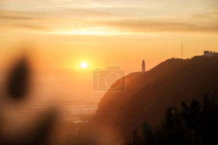 Téléchargez les photos : Coucher de soleil sur la côte avec phare debout sur la falaise au loin, océan, ciel orange et rétroéclairé à la grande route de l'océan, Victoria, Australie - en image libre de droit