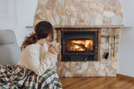 Photo for Young woman sitting at home by the fireplace with a hot tea or coffee mug and warming her hands, she is wearing white woollen sweater. Cold houses in Europe concept during energy and gas crisis. - Royalty Free Image