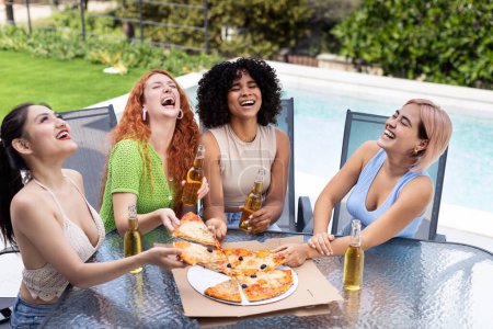 Foto de Cuatro niñas alegres de diferentes etnias se ríen, comen pizza y beben cerveza. El ambiente de celebración, felicidad y amistad. Mujeres chinas, afroamericanas, latinas e hispanas están juntas. - Imagen libre de derechos