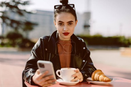 Téléchargez les photos : Belle jeune femme brune assise dans un café de rue avec un café et un croissant, elle prend un petit déjeuner ou juste une pause, se prélassant aux rayons du soleil. Elle tient un téléphone dans ses mains - en image libre de droit