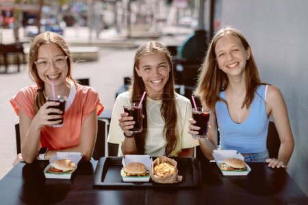 Foto de Tres adolescentes sonrientes y alegres (14-15 años) sentadas en un café al aire libre, disfrutando de hamburguesas y bebidas frescas de cola. Señoritas sosteniendo copas de coca, listas para beberlas con placer. - Imagen libre de derechos