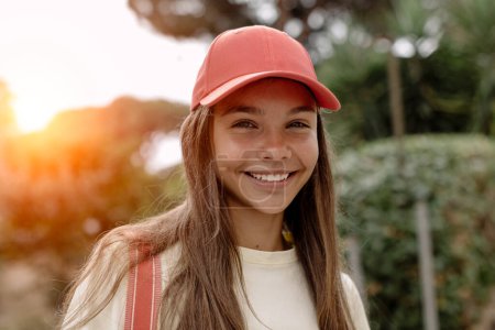 Téléchargez les photos : Portrait d'une belle adolescente (14-15 ans) coiffée d'une casquette de baseball rose, debout à l'extérieur, souriant doucement sur fond de coucher de soleil. - en image libre de droit