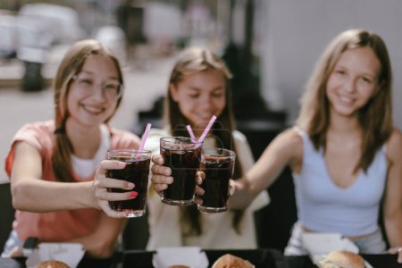 Téléchargez les photos : Trois adolescentes joyeuses et souriantes (14-15 ans) assises dans un café à l'extérieur, dégustant des boissons fraîches au cola. Des jeunes femmes qui prennent des verres à coke, prêtes à les boire avec plaisir. Focus sur les lunettes - en image libre de droit