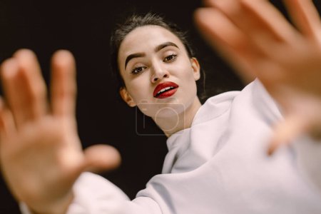 Photo for A stunning young woman, viewed from below, gazes confidently into the camera. Her lips adorned with red lipstick, her hands frame her face. Concept of modern femininity, beauty and gen Z culture. - Royalty Free Image
