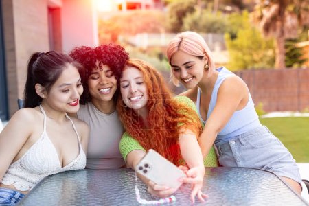 Un grupo diverso de mujeres jóvenes hermosas de varias nacionalidades se sienta en una mesa, tomando un selfie del atardecer en un solo teléfono. La imagen encarna la amistad intercultural, la diversión veraniega y la unión