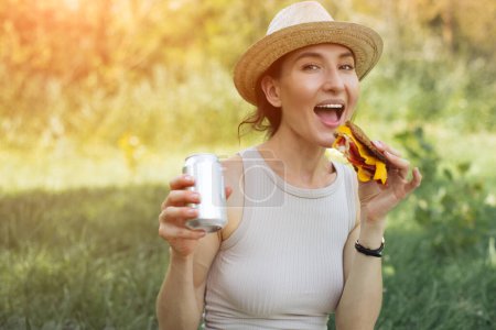 Foto de Retrato de una hermosa joven sonriente con un sándwich (jamón y queso) en una mano y una cerveza en la otra, disfruta de un picnic en un prado verde o bosque, abrazando el concepto de relajación al aire libre - Imagen libre de derechos