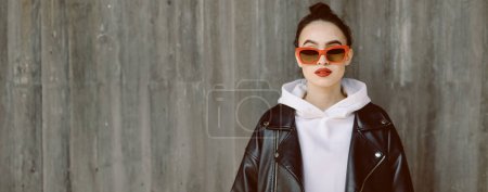 Photo for A beautiful young woman stands against a grey wall on the street, wearing red-framed glasses and red lipstick. Modern generation Z girl isolated portrait, image with copy space. - Royalty Free Image