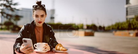 Téléchargez les photos : Belle jeune femme brune assise dans un café de rue avec un café et un croissant, elle prend un petit déjeuner. Elle tient un smartphone dans ses mains. Image de bannière panoramique avec espace de copie. - en image libre de droit