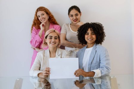 Photo for Multiethnic group of young ladies office workers or businesswomen are holding white blank paper and smiling. Diverse women are sitting at the table together, concept of international collaboration. - Royalty Free Image