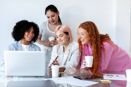Foto de Cuatro chicas sonrientes, enfocadas y creativas, se reunieron alrededor de una computadora, exudando un ambiente de trabajo profesional y colaborativo. Grupo multirracial de mujeres jóvenes que trabajan o estudian junto con pc. - Imagen libre de derechos