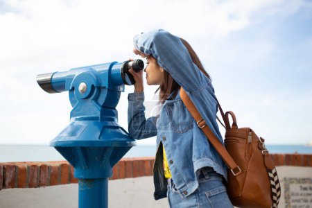 Téléchargez les photos : Vue latérale d'une jeune touriste utilisant un télescope pour voir l'océan - en image libre de droit