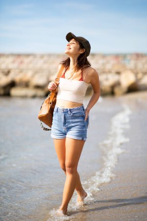Téléchargez les photos : Portrait complet du corps de jolie jeune femme avec casquette marchant sur la plage avec sac en cuir - en image libre de droit