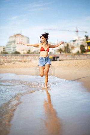 Photo for Full length portrait of carefree young woman running on the beach with arms outstretched - Royalty Free Image