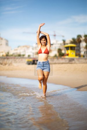 Foto de Retrato de cuerpo completo de una joven feliz en bikini que corre por la playa con los brazos levantados - Imagen libre de derechos