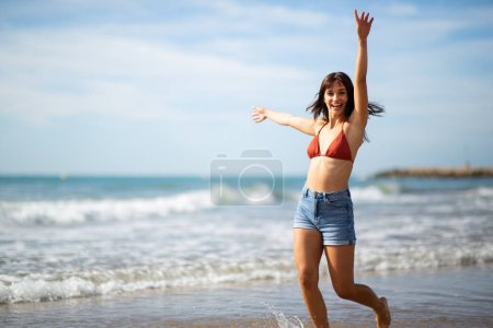 Photo for Portrait of cheerful young woman in bikini top running on the beach with arms raised - Royalty Free Image