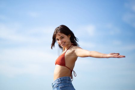 Téléchargez les photos : Portrait de jolie jeune femme debout avec les bras tendus contre le ciel regardant en arrière à la caméra - en image libre de droit