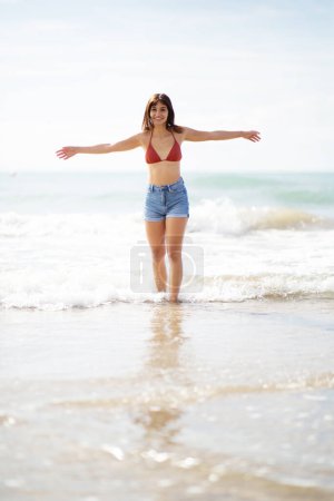 Photo for Full length portrait of happy young woman walking in sea water on the beach with arms outstretched - Royalty Free Image