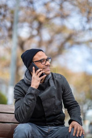 Photo for Portrait of smiling young african man in winter jacket and hat enjoying talking on mobile phone sitting on bench outside - Royalty Free Image