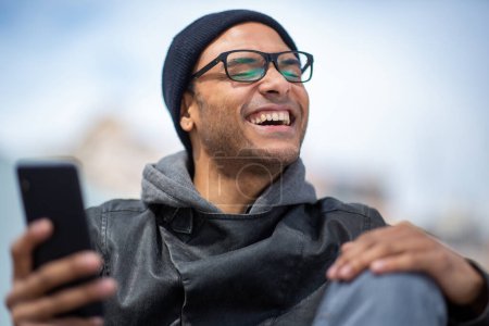 Photo for Close up portrait of black man sitting outside in the city with mobile phone and smiling - Royalty Free Image