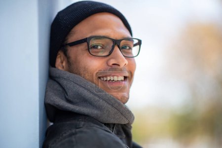 Foto de De cerca retrato de hombre afroamericano guapo con gafas inclinadas a una pared y mirando a la cámara sonriendo - Imagen libre de derechos