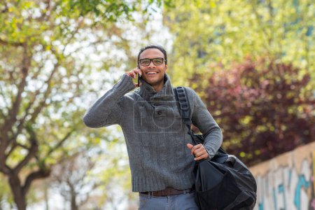 Foto de Retrato de un joven sonriente con una bolsa caminando afuera y hablando por teléfono móvil - Imagen libre de derechos
