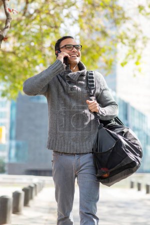 Photo for Portrait of stylish young man talking on mobile phone carrying bag in the city - Royalty Free Image