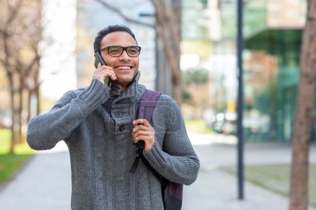Foto de Retrato de un atractivo joven africano con una bolsa caminando afuera y hablando por teléfono - Imagen libre de derechos