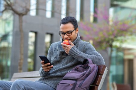 Foto de Retrato de un joven africano comiendo una manzana y usando un teléfono celular mientras está sentado en el banco exterior de la ciudad - Imagen libre de derechos