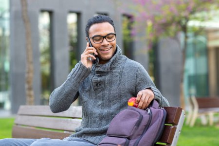 Photo for Portrait of happy young black man talking on cell phone while sitting on bench with bag outside in city - Royalty Free Image