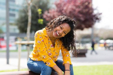 Téléchargez les photos : Portrait d'une jeune femme africaine séduisante riant alors qu'elle était assise dehors dans la ville - en image libre de droit