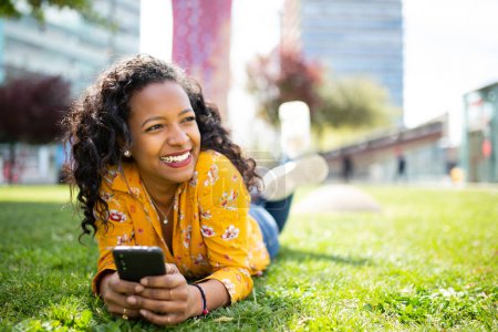 Foto de Retrato de una joven negra sonriente tumbada en la hierba con un teléfono móvil mirando hacia otro lado - Imagen libre de derechos