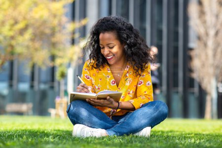 Photo for Portrait of beautiful young african woman sitting on grass outside and writing in book - Royalty Free Image