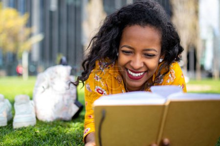 Foto de Retrato de cerca de una joven africana sonriente tumbada en el césped en el parque leyendo un libro - Imagen libre de derechos