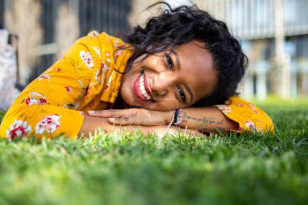 Téléchargez les photos : Gros plan portrait de belle jeune femme africaine allongée sur l'herbe regardant la caméra et souriant - en image libre de droit