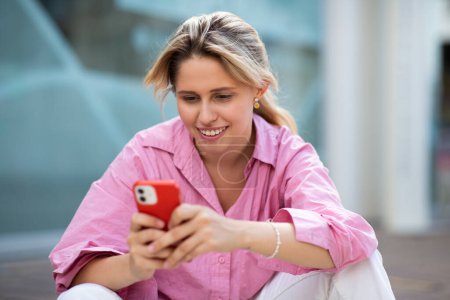 Téléchargez les photos : Portrait de belle jeune femme utilisant un téléphone portable tout en étant assis à l'extérieur dans la ville - en image libre de droit