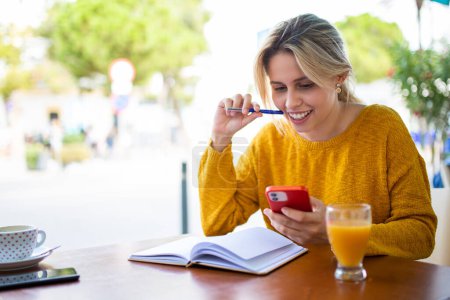 Téléchargez les photos : Portrait de belle jeune femme assise au café extérieur tenant stylo regardant téléphone portable avec livre sur la table - en image libre de droit