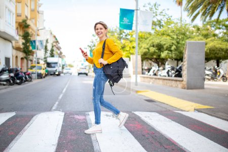 Photo for Full length portrait of cheerful young latin woman with handbag and cellphone crossing a street - Royalty Free Image