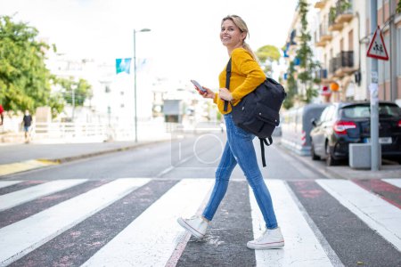 Photo for Full length portrait of pretty young latin woman walking on zebra crossing with handbag and cellphone - Royalty Free Image