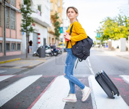 Photo for Full body portrait of young woman traveller crossing the street with suitcase and handbag - Royalty Free Image