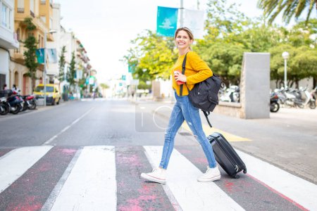 Photo for Full body portrait of happy young woman crossing the street with travel bag - Royalty Free Image