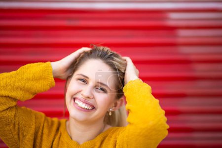Foto de Retrato de cerca de una joven alegre sonriendo con las manos en la cabeza contra el obturador rojo afuera - Imagen libre de derechos