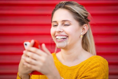 Téléchargez les photos : Gros plan portrait d'une jeune femme souriante regardant un téléphone portable contre un volet rouge à l'extérieur - en image libre de droit