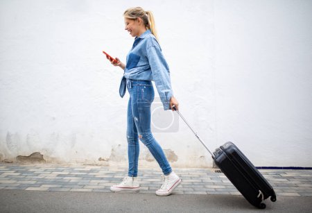 Foto de Retrato de vista lateral de mujer joven caminando con bolsa de viaje y usando teléfono móvil - Imagen libre de derechos