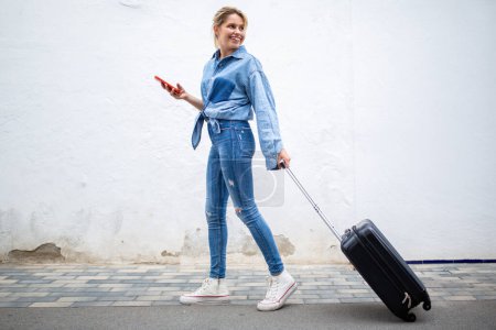 Téléchargez les photos : Portrait corporel complet de la jeune femme heureuse marchant avec sac à valise et téléphone portable regardant derrière - en image libre de droit