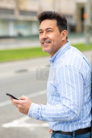 Téléchargez les photos : Portrait latéral de l'homme mûr marchant à l'extérieur dans la ville avec téléphone mobile - en image libre de droit