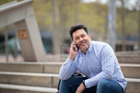 Photo for Portrait of happy mature man sitting outdoors on steps talking on cell phone - Royalty Free Image