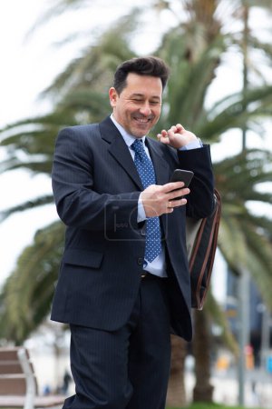 Foto de Retrato de hombre de negocios maduro feliz con bolsa de traje y el uso de su teléfono celular caminando fuera - Imagen libre de derechos